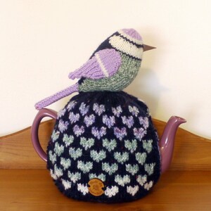 Könnte beinhalten: Eine gestrickte Vogel-Teekanne mit Herzmuster in Lila und Grün. Die Teekanne ist auf einer lila Teekanne mit einem Ausguss.