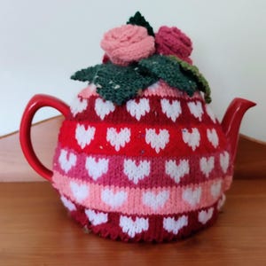 Könnte beinhalten: Eine rote Teekanne mit einer gestrickten Teekanne, die ein Herzmuster in Rosa und Weiß aufweist. Die Teekanne ist mit einer Gruppe von rosa gehäkelten Blumen und grünen Blättern verziert.