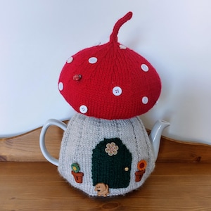 May include: A knitted teapot cosy shaped like a mushroom house. The mushroom cap is red with white polka dots and the house is grey with a green door and a small brown dog figurine.