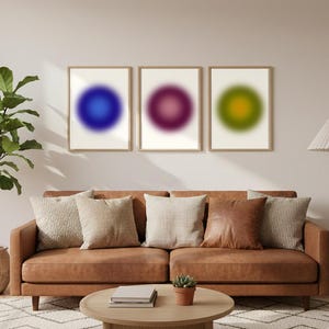 Puede incluir: Tres impresiones artísticas enmarcadas con diseños circulares borrosos en tonos azules, morados y verdes. La obra de arte cuelga sobre un sofá de cuero marrón con cojines decorativos. Una mesa de centro redonda de madera está frente al sofá.