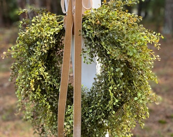 May include: A green leafy wreath with a brown velvet ribbon tied around it. The wreath is hanging on a white post.