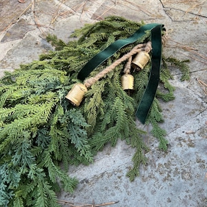 May include: A festive evergreen garland with a dark green velvet ribbon and three small gold bells. The garland is made of faux fir branches in varying shades of green and blue-green. A rustic holiday decoration.