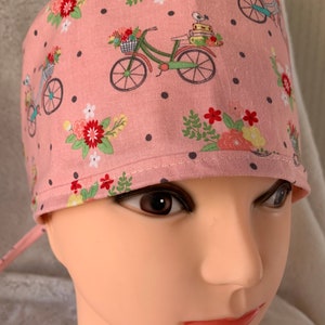 May include: Pink surgical scrub hat with a repeating pattern of bicycles with flowers, birds, and books. The hat has black polka dots and a tie at the back.