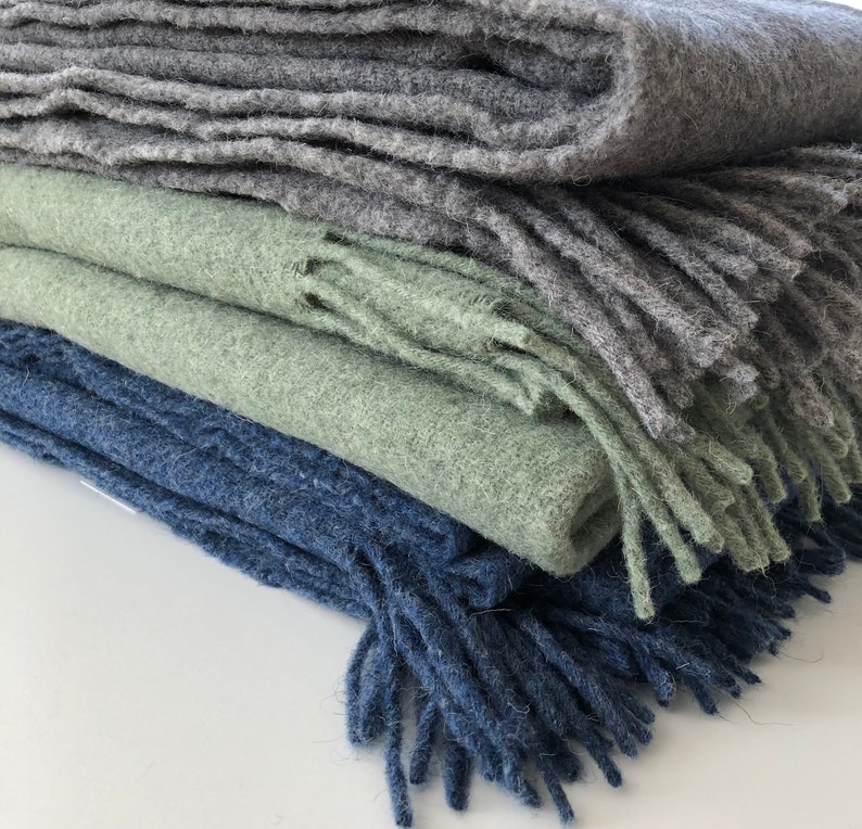 May include: A stack of three blankets in different shades of blue, green, and gray. The blankets are made of a soft, fuzzy material and have fringe edges.