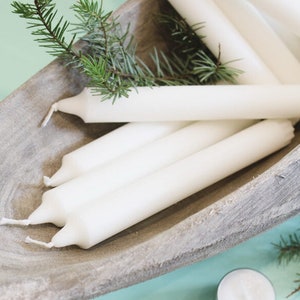 Op de afbeelding: Een close-up van een houten kom gevuld met witte kaarsen. De kaarsen zijn in een stapel gerangschikt, met de punten van de kaarsen die in verschillende richtingen wijzen. De kom is gemaakt van licht hout met een ruwe textuur.