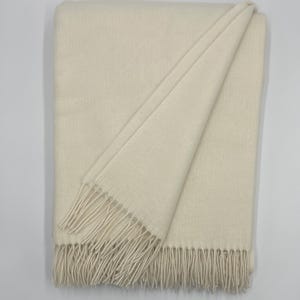 May include: Cream-colored blanket with a soft, textured appearance. The blanket is folded, with a portion draped over, revealing a fringe detail along the edge. The neutral color and simple design suggest a cozy home textile.