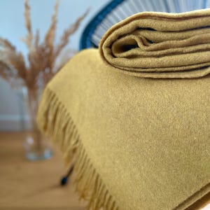 Könnte beinhalten: Senffarbene Decke mit einem gefalteten Abschnitt oben. Die Decke hat eine weiche Textur und einen Fransenrand. Der Hintergrund umfasst einen verschwommenen Stuhl und eine Vase mit getrockneten Pflanzen.