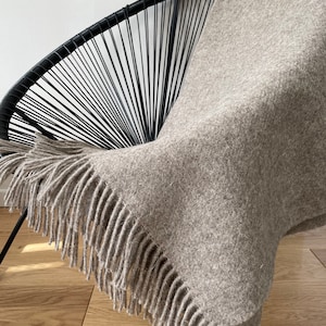 May include: A light brown throw blanket with fringe on all sides draped over a black wire chair. The blanket is made of a soft, fuzzy material.