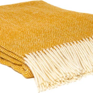Könnte beinhalten: Senfgelbe Decke mit Fischgrätenmuster. Die Decke hat einen cremefarbenen Fransenbesatz an einer Kante. Die gefaltete Decke besteht aus einem weichen Material und eignet sich für die Inneneinrichtung.