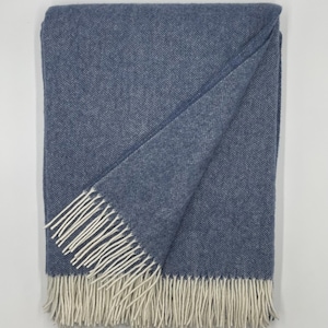 May include: A folded blue throw blanket featuring a classic herringbone pattern throughout the fabric. The blanket is finished with long, cream-colored fringe tassels along the bottom edge, providing a textured contrast to the woven blue textile.