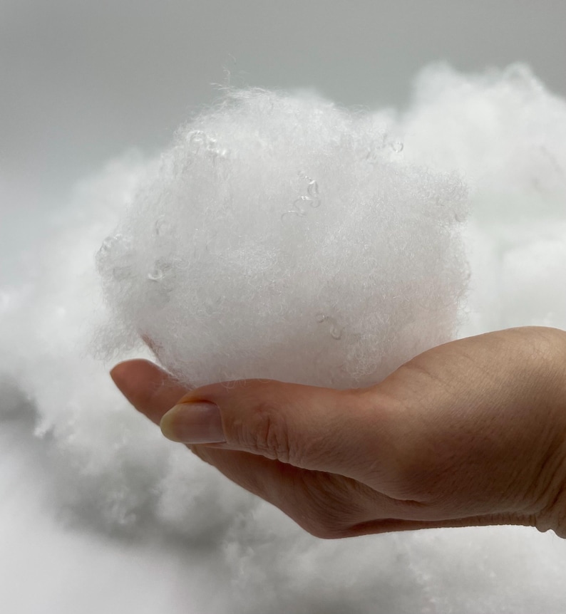 May include: A close-up of a hand holding a large clump of white fibrefill. The fibrefill is soft and fluffy, and it looks like it could be used for stuffing pillows or other crafts.