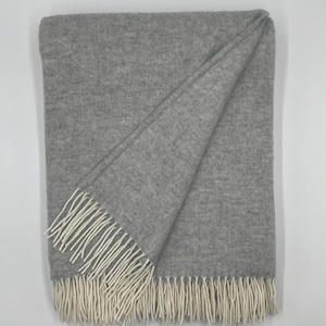 Puede incluir: Una manta rectangular de color gris con un sutil patrón de tejido en espiga. La manta está doblada en una esquina para revelar la textura y está terminada con largos flecos de color crema a lo largo del borde inferior.