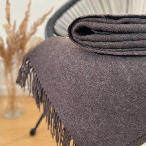 Op de afbeelding: Een opgevouwen, donkergrijze deken met een gefranjerde rand. De deken is over een stoel gedrapeerd, met een deel opgevouwen. De zachte, getextureerde stof is effen van kleur en de franje heeft een bijpassende grijstint.