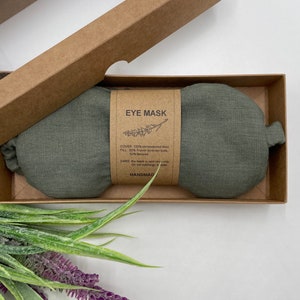 Pu&ograve; includere: Una maschera per gli occhi in lino verde riempita con boccioli di lavanda francese e semi di lino. L'etichetta recita "EYE MASK" e include le istruzioni per la cura. Fatto a mano.