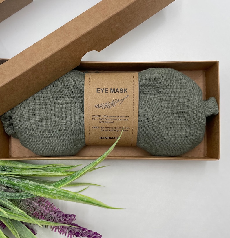 Pu&ograve; includere: Una maschera per gli occhi in lino verde riempita con boccioli di lavanda francese e semi di lino. L'etichetta recita "EYE MASK" e include le istruzioni per la cura. Fatto a mano.