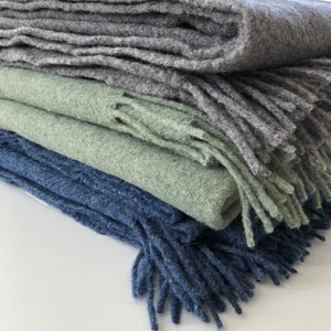 May include: A stack of three blankets in different shades of blue, green, and gray. The blankets are made of a soft, fuzzy material and have fringe edges.