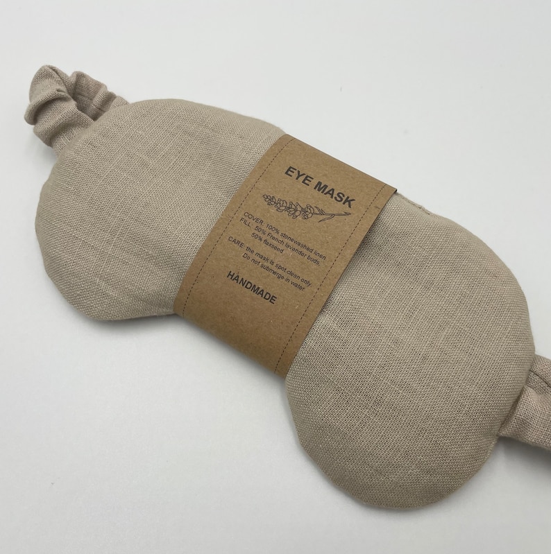 Może przedstawiać: Beżowa lniana maska na oczy z elastyczną opaską. Brązowa papierowa etykieta z napisem "EYE MASK" zawiera listę materiał&oacute;w: kamieniowany len, francuskie pąki lawendy i siemię lniane. Na etykiecie widnieje r&oacute;wnież informacja, że maska jest ręcznie robiona.