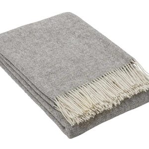 Puede incluir: Una manta rectangular doblada que presenta un patrón de espiga de color gris claro en toda la tela. La manta está terminada con una hilera de flecos de color crema a lo largo de uno de los bordes más cortos, añadiendo textura al diseño.