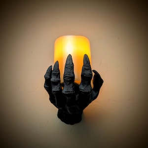 Puede incluir: Una lámpara de pared de mano esquelética negra con una luz cálida y brillante que emana del portavelas.