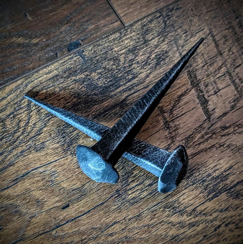 May include: Two black metal nails with pointed tips, lying on a wooden surface.