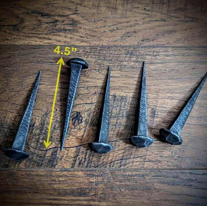 May include: Six black metal nails with pointed tips, arranged in a row on a wooden surface. The nail on the left is 4.5 inches long.