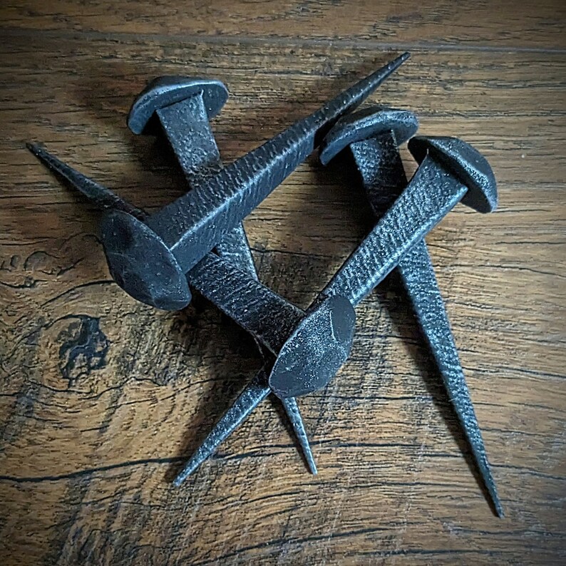May include: Three black metal nails with pointed tips, arranged in a criss-cross pattern on a wooden surface.