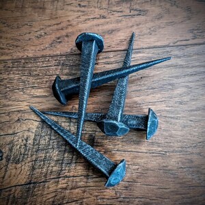 May include: Four black metal nails with pointed tips and flat heads. The nails are arranged in a cluster on a wooden surface.