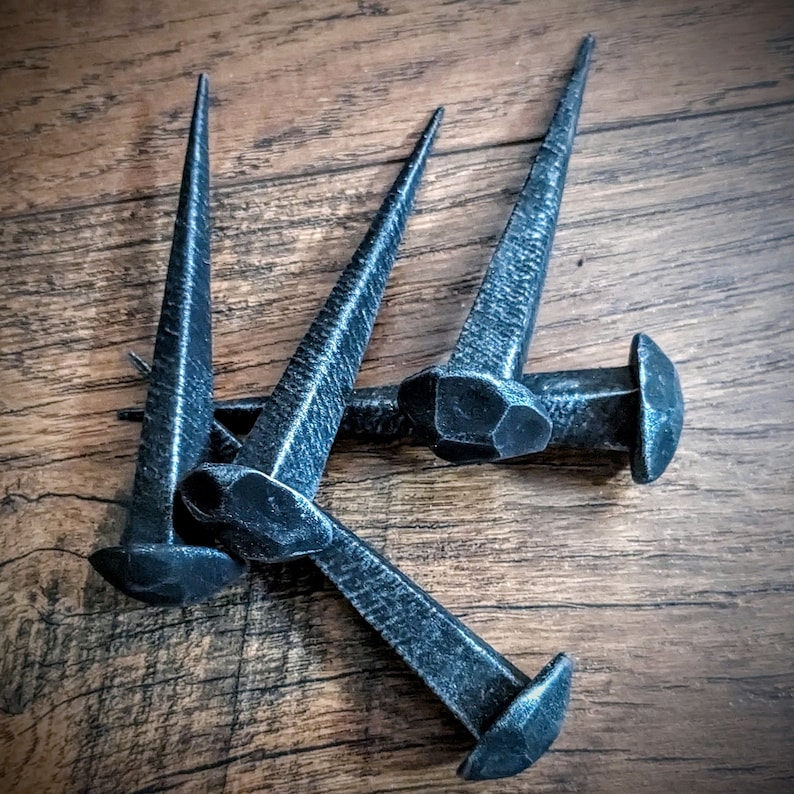 May include: Three large, hand-forged iron nails with blunt, square heads and pointed tips. The nails are arranged in a cluster on a wooden surface.