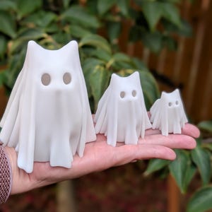May include: Three white ghost figurines of varying sizes. Each ghost has a draped sheet with two eye holes. The figurines are held in a hand, with a green leafy background. The ghosts have a cat-like appearance.