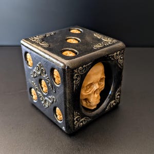 May include: A black and gold decorative die with skull and scrollwork details. The die has six sides, each with a different number of skull-shaped pips. A unique gothic-inspired item.