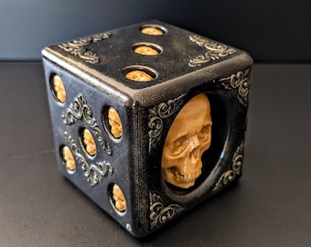 May include: A black and gold decorative die with skull and scrollwork details. The die has six sides, each with a different number of skull-shaped pips. The die is a unique and gothic-inspired item.