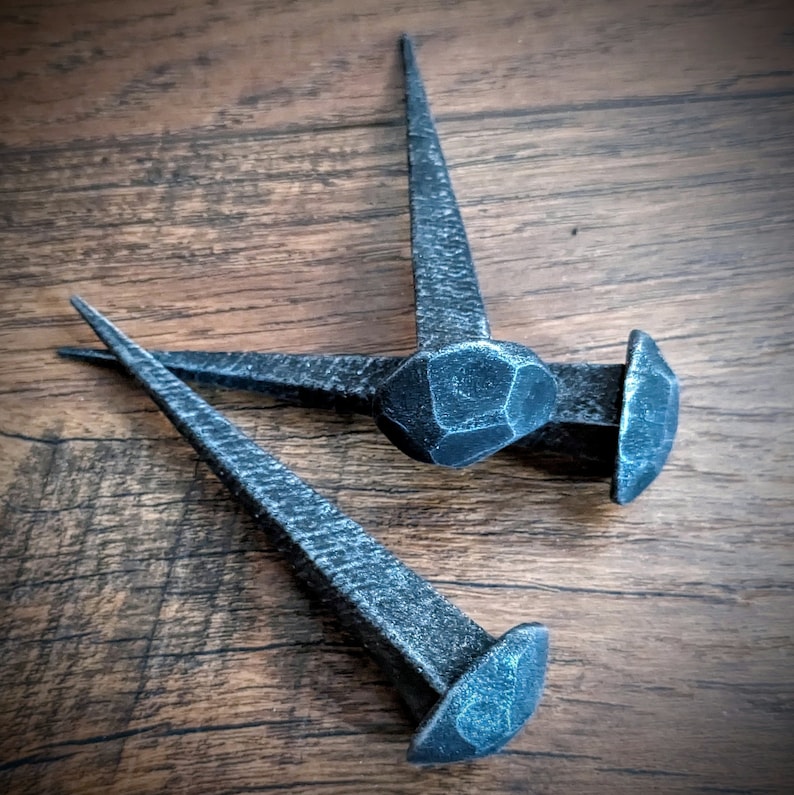 May include: Three antique-style, hand-forged iron nails with hammered heads. The nails are arranged in a triangular shape on a wooden surface.