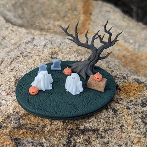 May include: A miniature Halloween scene with a bare tree, two white ghosts, three gray tombstones, and three orange pumpkins on a green base.