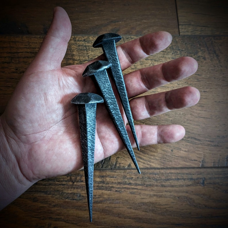 May include: Three black metal nails with pointed tips, arranged in a hand. The nails are approximately 10 cm long.