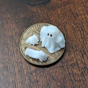 May include: A set of four white plastic Halloween decorations, including a ghost with two black eyes, a small ghost, and two bone-shaped pieces. The decorations are sitting on top of a US penny.