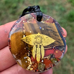 Könnte beinhalten: Ein runder Anhänger aus Kunstharz mit eingebetteten Tigeraugen-Steinfragmenten, roten Jaspisstücken und goldenem Glitzer. Ein goldfarbener Metallanhänger in Form eines Traumfängers mit drei hängenden Federn ist mittig im Harz platziert.