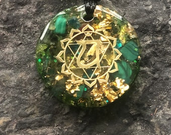 May include: A round pendant necklace with a black cord. The pendant is a clear resin with green stones and gold flakes. A gold-colored symbol is in the center of the pendant.