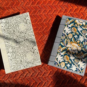 May include: Two hardcover journals with different patterned covers. The journal on the left has a white background with a black and gray floral pattern. The journal on the right has a dark blue background with a white and yellow floral pattern.