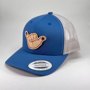 May include: A blue and white trucker hat with a brown wooden hand gesture patch. The patch is a shaka sign, a Hawaiian gesture of peace and good vibes. The hat has a black label that reads "The Authentic Yupoong Snapback Yupoong Since 1974".