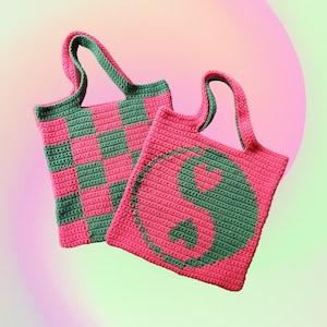 May include: Two handmade crochet tote bags. One bag features a pink and green checkerboard pattern. The other bag has a pink background with a green yin-yang symbol and a heart. Both bags have matching green handles.