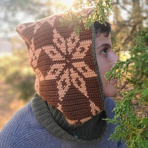 May include: A brown and peach crocheted hooded cowl with a snowflake pattern. The cowl is worn by a person with brown hair and is partially obscured by green foliage.