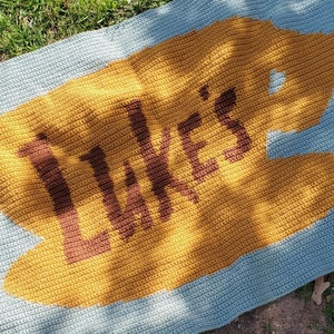 Pode incluir: Um cobertor de bebé em crochê azul e amarelo com o nome "Luke's" em letras castanhas.