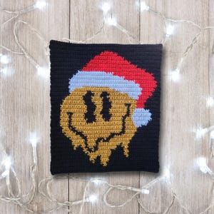 May include: A crocheted square pillow with a yellow smiley face wearing a red and white Santa hat. The pillow is on a wood background with white Christmas lights.