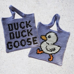Może przedstawiać: Dwie fioletowe torby na zakupy wykonane na szydełku. Jedna torba ma napis "Duck Duck Goose" czarnymi literami. Druga torba ma białego, szydełkowego kaczora z pomarańczowymi łapkami i żółtym dziobem.