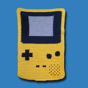 May include: A handmade crochet blanket in the shape of a retro handheld video game console. The blanket features a yellow body with a navy blue screen border and a light blue screen. Black buttons and a directional pad are also visible.