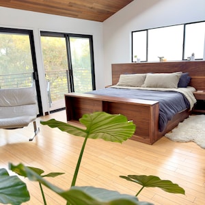 Könnte beinhalten: Ein modernes Schlafzimmer mit einem Bettrahmen, Kopfteil und Nachttisch aus Holz. Das Bett ist mit blauer und beiger Bettwäsche bezogen. Der Raum hat eine Holzdecke und einen Holzboden, mit großen Fenstern und einem Oberlicht. Ein weißer Ledersessel ist sichtbar.