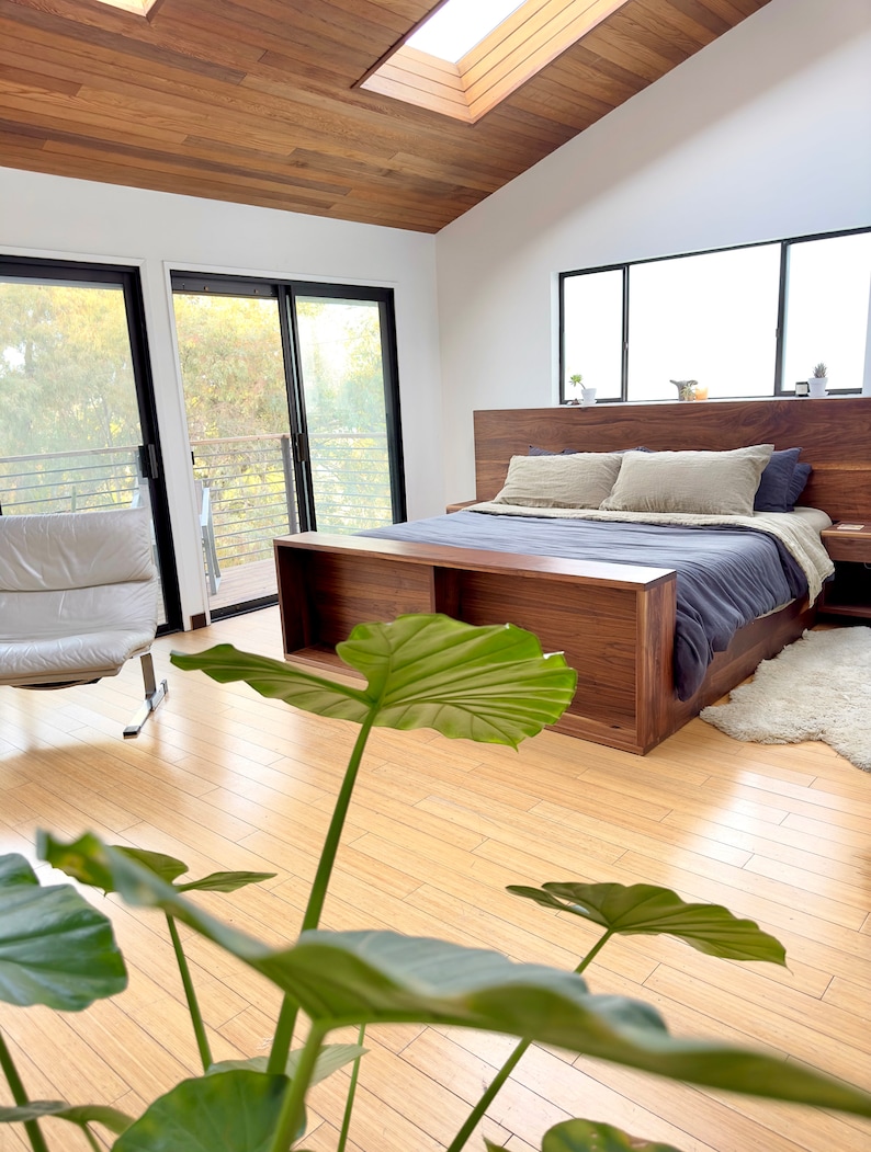 May include: A modern bedroom with a wooden bed frame, headboard, and nightstand. The bed is made with blue and beige bedding. The room has a wooden ceiling and floor, with large windows and a skylight. A white leather chair is visible.