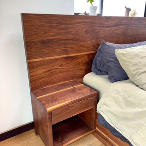 May include: A dark brown wooden nightstand with a drawer and open shelf, attached to a matching headboard. The headboard and nightstand have a natural wood grain finish. The bed is made up with blue and beige bedding.