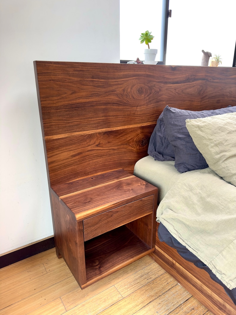 May include: A dark brown wooden nightstand with a drawer and open shelf, attached to a matching headboard. The headboard and nightstand have a natural wood grain finish. The bed is made up with blue and beige bedding.