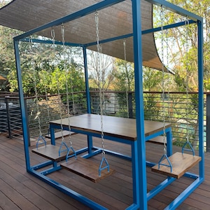 May include: A blue metal outdoor dining set with a rectangular table and four attached swings. The table is made of wood and is surrounded by a blue metal frame. A canopy provides shade. The set is on a wooden deck.